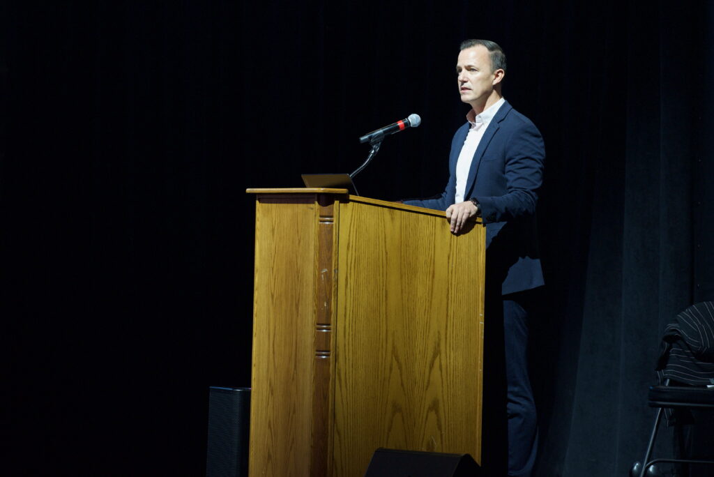 Jay Renkels with MIG stands in a suit at a podium while delivering a keynote address to a crowd of hundreds at Ed Robson Arena.