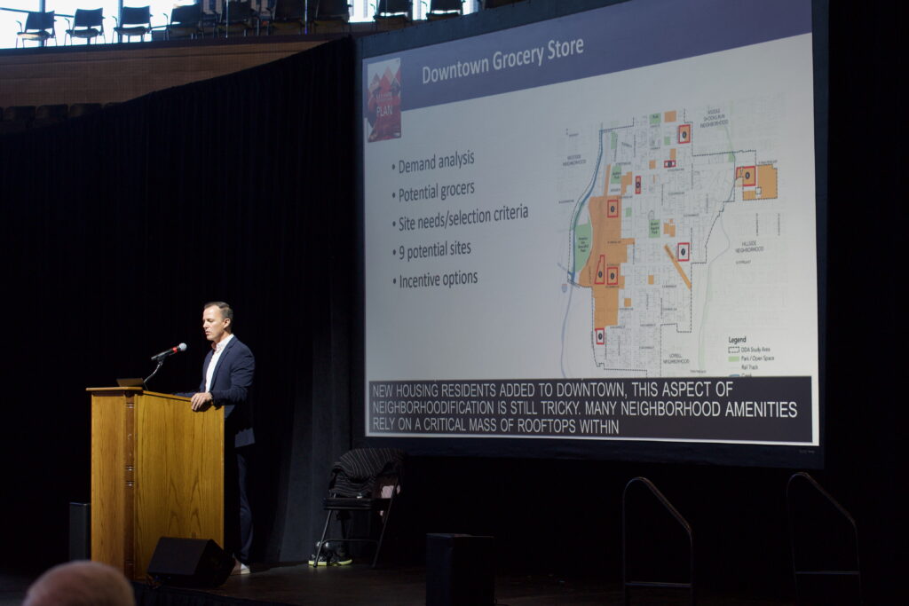 Jay Renkels with MIG stands in a suit at a podium while delivering a keynote address to a crowd of hundreds at Ed Robson Arena.
