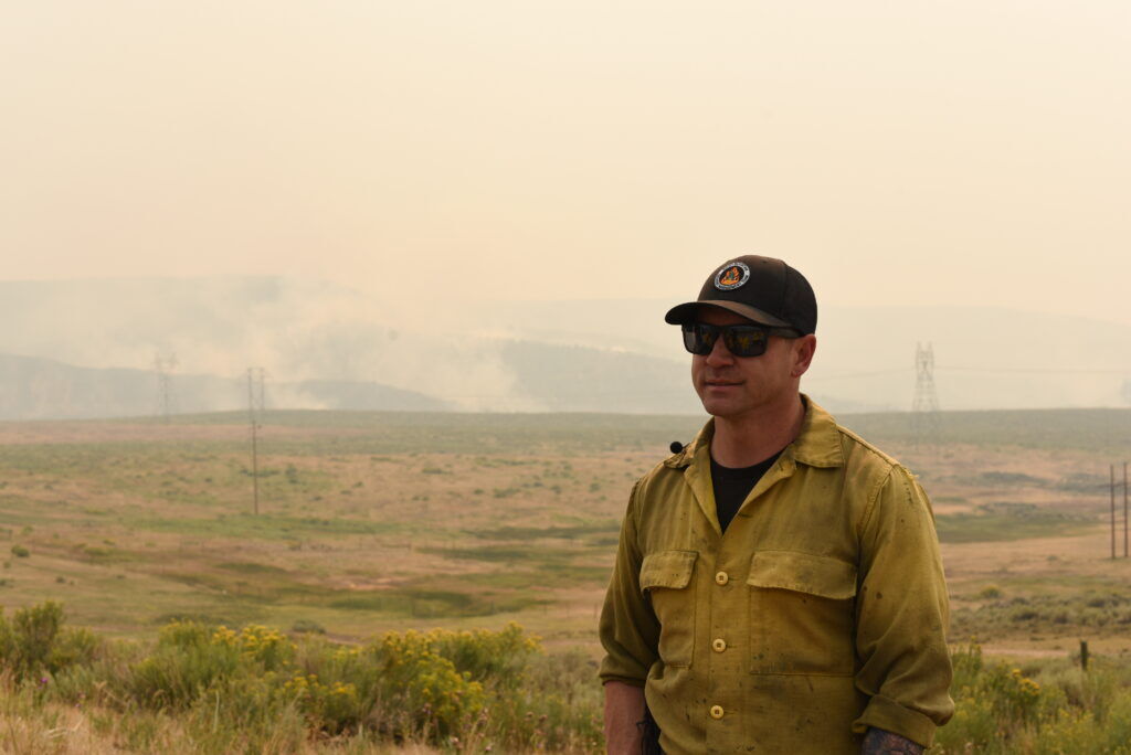 A man stands in front of a fire burning behind some short grass