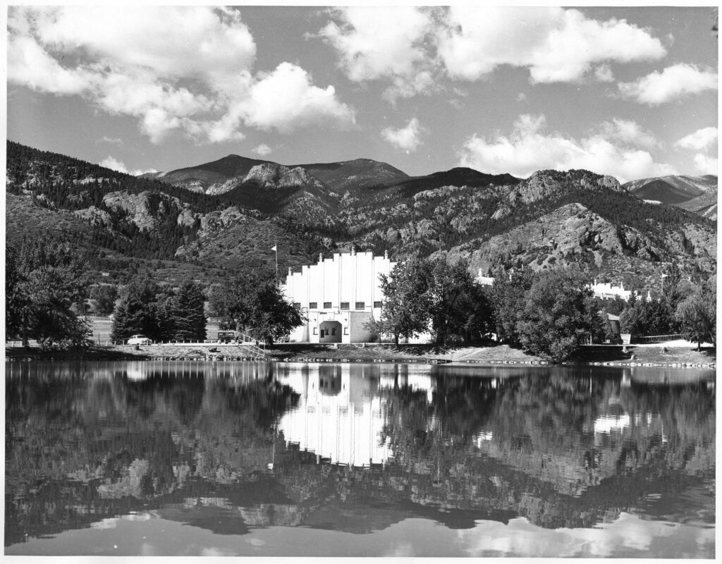 Long view of the Broadmoor Ice Arena [first Broadmoor World Arena] across Broadmoor Lake, with the building and mountains behind reflected in the water. Two automobiles from the late 1940s and the early 1930s are parked on the left. Research indicates that the first Broadmoor World Arena was a skating rink and hockey arena located at the Broadmoor Resort. Originally an outdoor equestrian center and riding academy, a building was constructed over the area and converted into an ice arena which opened in January 1938. Photo Courtesy Margaretta M Boas photo collection Regional History & Genealogy, Pikes Peak Library District 001-3553
