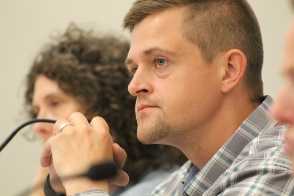 Close-up of Hudson Town Manager Bryce Lange seated at the dais during a town board meeting. He listens attentively with his hands clasped near a microphone as public comment is underway.