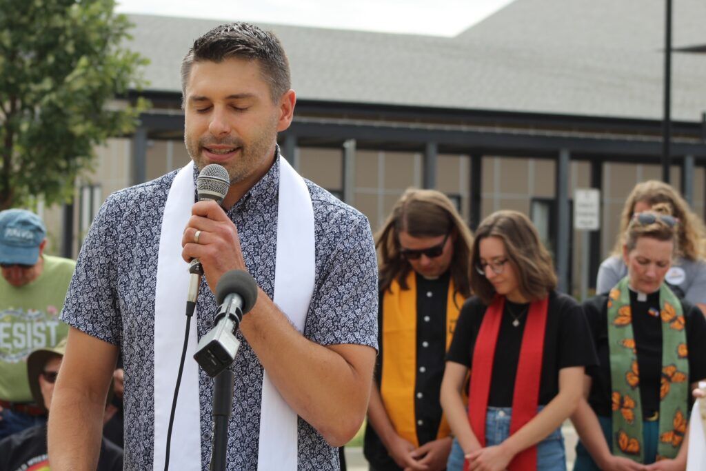 A man with a microphone leads a prayer in front of group with bowed heads.