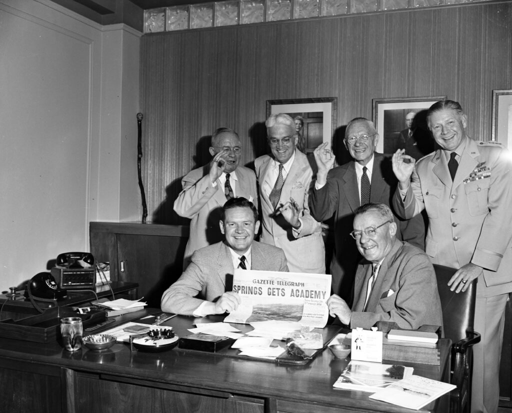 Five civilian civic leaders and a four-star U.S. Air Force general officer celebrate the selection of Colorado Springs as the site for the U.S. Air Force Academy. The seated men display the headlines of the Colorado Springs Gazette Telegraph newspaper that states "Springs Gets Academy." Identified in Gazette article on 6/24/1954 as: seated (from left) Thayer Tutt and Russell Law, co-chairmen of the Chamber of Commerce Military Affairs Committee. In back (from left) Charles Tutt, Chase Stone, Lt. Gen. O.W. Griswold (ret.) and Gen. Benjamin Chidlaw of Air Defense Command. Photo by Stanley Payne Courtesy of The Gazette and Regional History & Genealogy, Pikes Peak Library District, 004-890