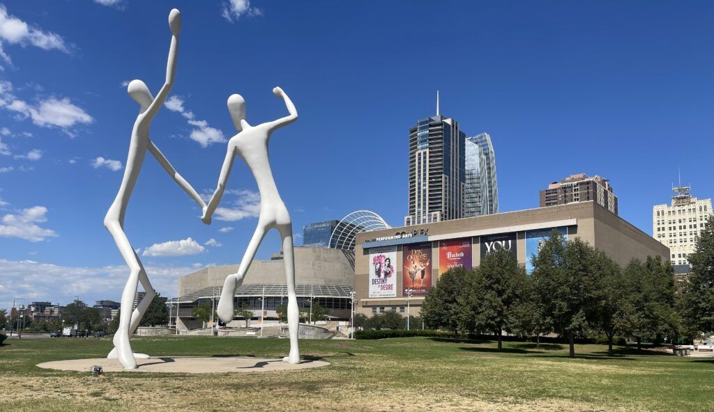 Two large white sculptures stand in Sculpture Park with Boettcher Concert Hall and the Denver Performing Arts Complex in the background under a blue sky.