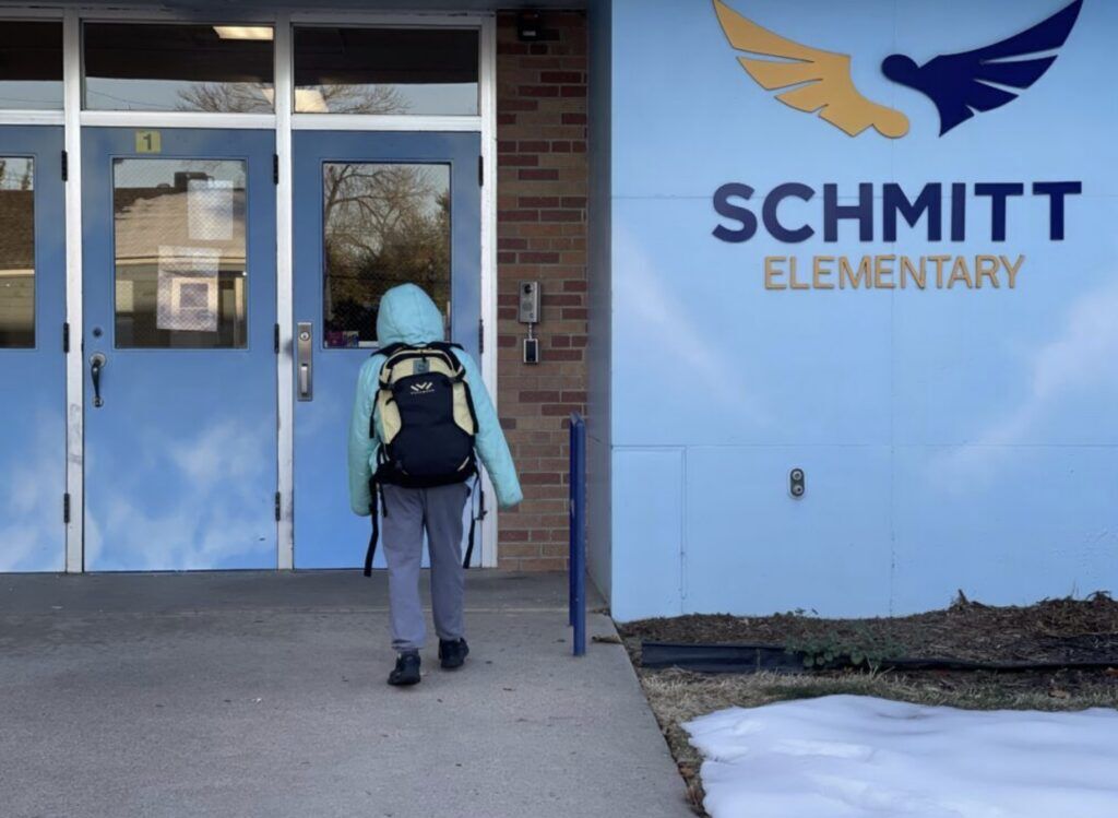 A student with a backpack approaches the blue entrance doors of Schmitt Elementary School.