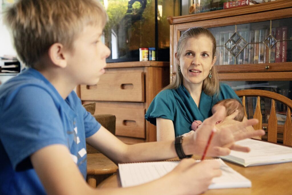 A woman holding a baby talks with a boy as he writes in a notebook at a dining room table.