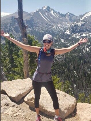 A smiling woman with arms spread stands on rocks with mountains in the background.