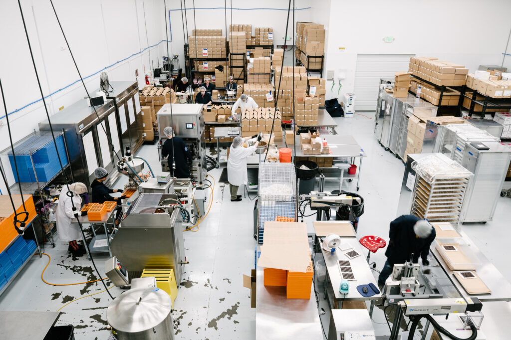 A high-angle view of the inside of a warehouse with various machines, cardboard boxes and people wearing lab coats and hairnets.
