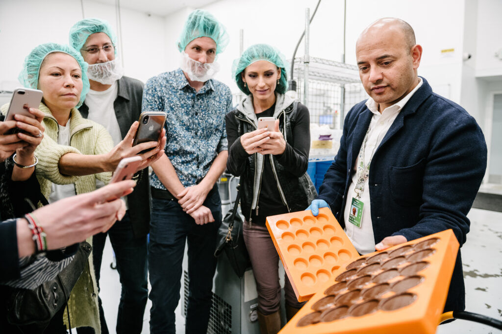 A bald man wearing a suit presents orange molds, one of which is filled with chocolate disks, to a group of people wearing hairnets and holding cellphones.