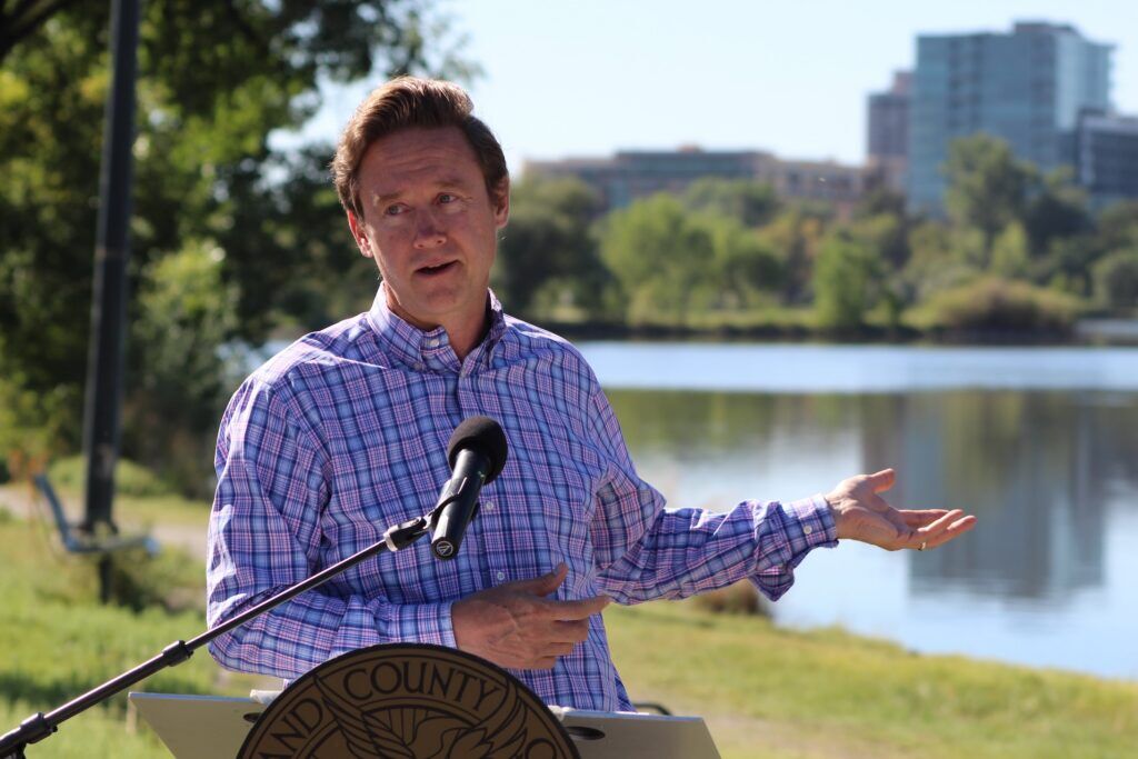 Denver Mayor Mike Johnston speaks at a podium with Sloan’s Lake in the background during an announcement about a $50 million lake restoration project.